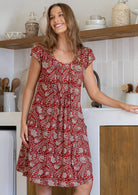 Woman wearing a 100% cotton rust red based floral dress with round neckline and pin tucks across bust. 
