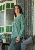 Woman stands sideways wearing a 100% cotton top showcasing the simple round neckline, long sleeves and pleats across the bodice.