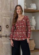 Woman wearing a red 100% cotton floral top standing in a room with wooden door and shelves.