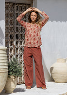 Woman wearing a red floral top and rust-colored pants standing in front of a textured wall and decorative pots.