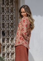 Woman standing sideways wearing a floral top and red pants standing next to a textured wall.