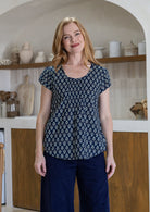 Woman wears navy blue based floral print 100% cotton cap sleeve top with pin tuck detail across the bust and a scoop neckline