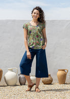 Woman wearing a moss green based floral  print short sleeve top with scoop round neckline and pleated detail across the bodice