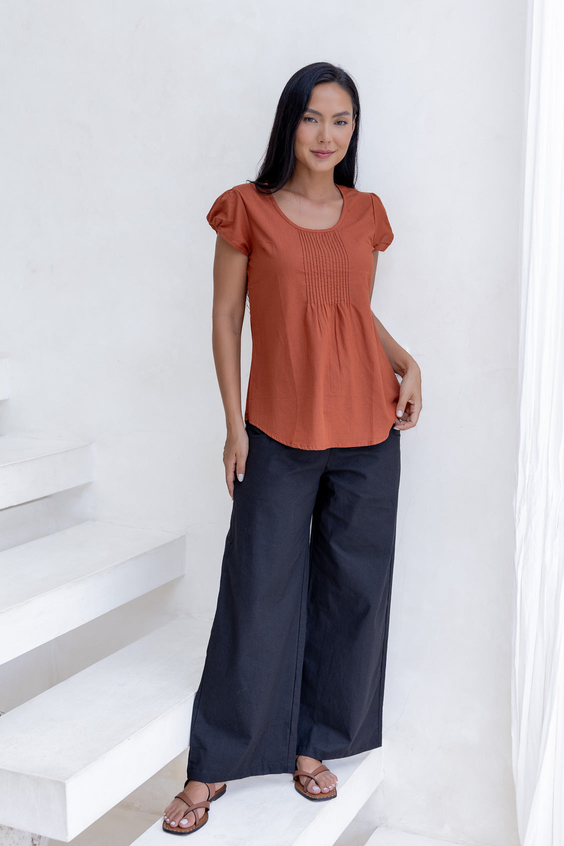 Woman wearing a rust orange cotton top with cap sleeves and pleated detailing, paired with dark cotton pants standing on a white staircase.