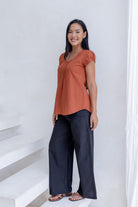 Woman poses in black cotton pants and relaxed fit cotton cap sleeved top in rust orange with pleated detailing across the bodice.