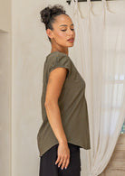 Woman in a green cap sleeved cotton top and black pants standing in a room with wooden shelves and a door.