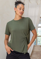 Woman wearing a soft stretch rayon T-shirt in olive green with a twist knot front, styled with black cotton pants hands in pockets 