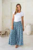 Woman wearing a cerulean blue based block style floral print pants with smocked elastic waistband.
