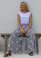 Woman sitting on a wooden bench wearing a white top and soft blue based floral wide leg pants with elastic waist.