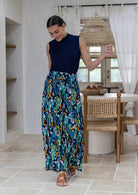A woman wearing a sleeveless navy blue mock-neck top and long, flowing, wide leg rayon pants in a colourful floral peacock print, and tan sandals