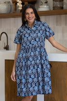 A woman wearing a short-sleeve buttoned bodice relaxed fit cotton dress with a blue, white, and light blue geometric pattern, standing in a kitchen setting.
