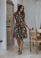 Model stands with one hand in pocket wearing a 100% cotton knee length shirt style retro floral dress in olive green based floral print.
