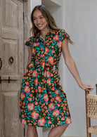 Woman in a olive green based floral cotton dress with cap sleeves, buttoned bodice, pockets, box pleats at the waist and a fabric waist tie.