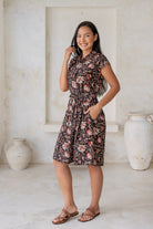 Woman in a black floral cotton dress with cap sleeves, buttoned bodice, pockets, box pleats at the waist and a fabric waist tie.