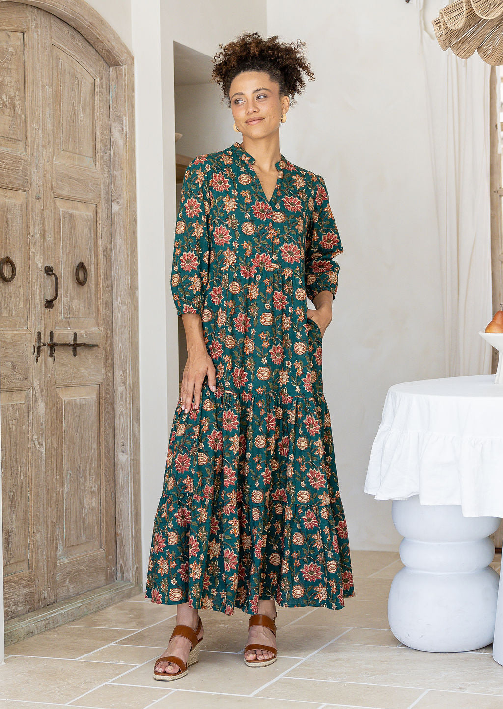 Woman wearing a green floral cotton dress standing in a room with wooden door and white table.