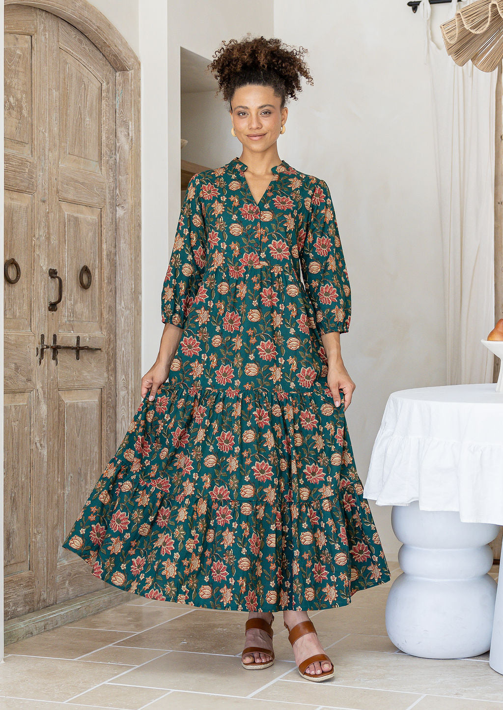 Woman wearing a green floral cotton dress with sleeves standing in a room with wooden door and white table.