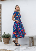 Woman stands sideways wearing a cotton button through cotton dress with hidden side pockets, short sleeves and a detachable waist tie.