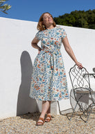 Woman wears retro style cotton button through midi length dress with high round neckline, short sleeves and a removable waist tie.
