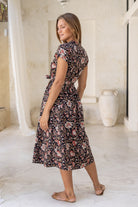 Model standing in back pose wearing a 100% cotton button through midi length dress with rust, tan and sand coloured florals on a black base.