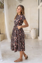 Model stands with hands in pocket wearing a retro style midi length cotton dress with button through front, fabric waist tie and short sleeves.
