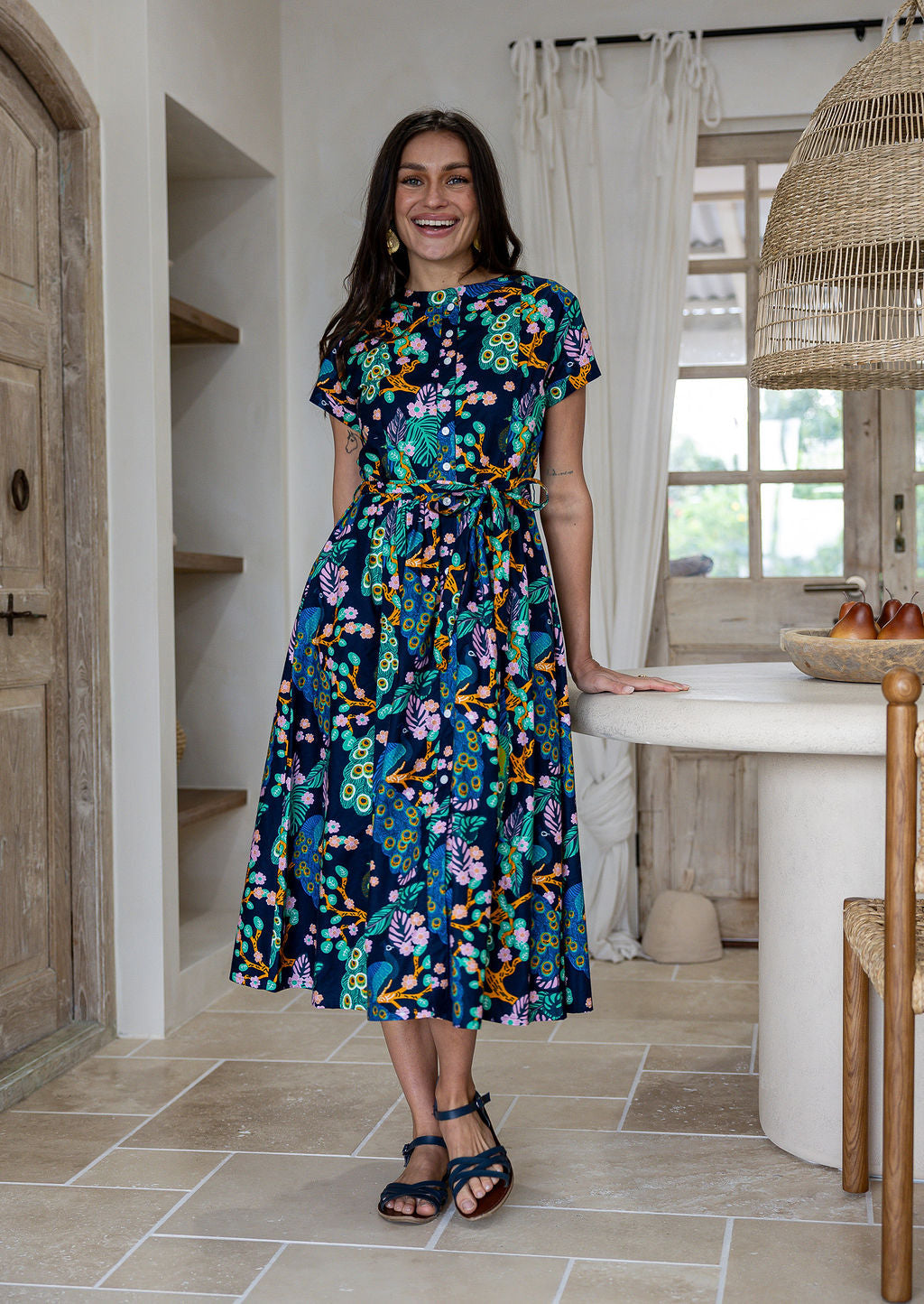 Woman wears retro style cotton button through midi length dress with high round neckline, short sleeves and a removable waist tie.