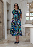 woman wearing a 50's style cotton button through midi length dress in navy based floral print with a removable waist tie and both hand in pockets