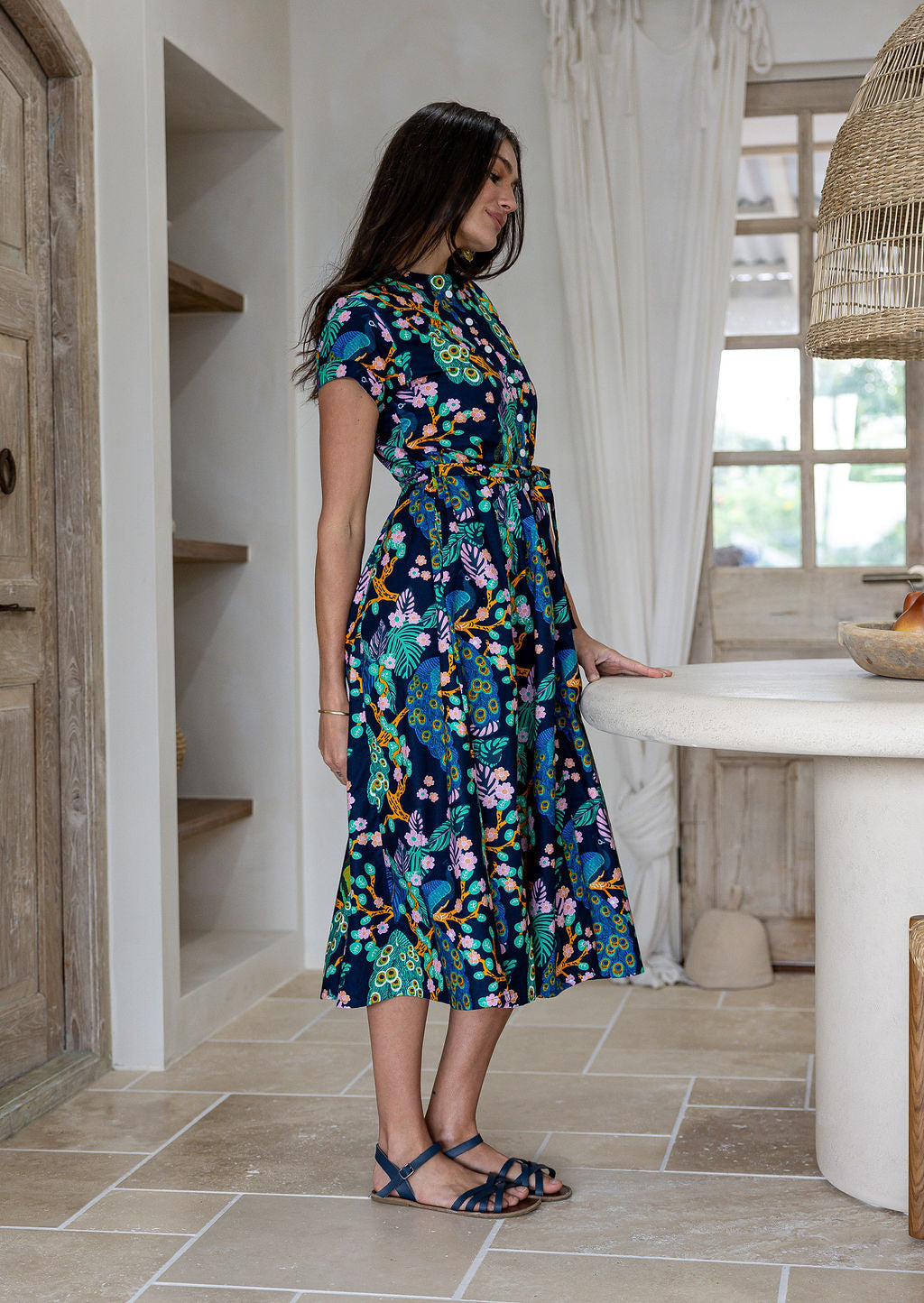 Woman standing sideways wearing a 1950's style short sleeved, button through cotton dress and high round neckline.