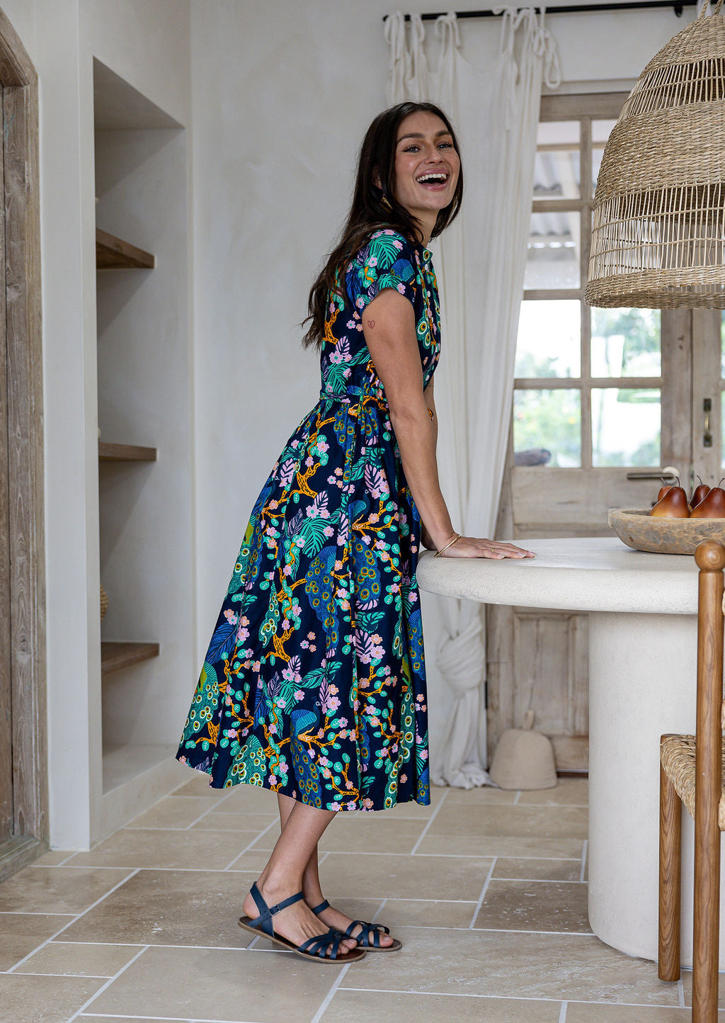 A woman wearing a navy blue midi dress with a colorful floral print, standing in a bright, modern kitchen setting.