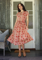 Woman wearing a floral dress standing in a room with wicker furniture and patterned walls.