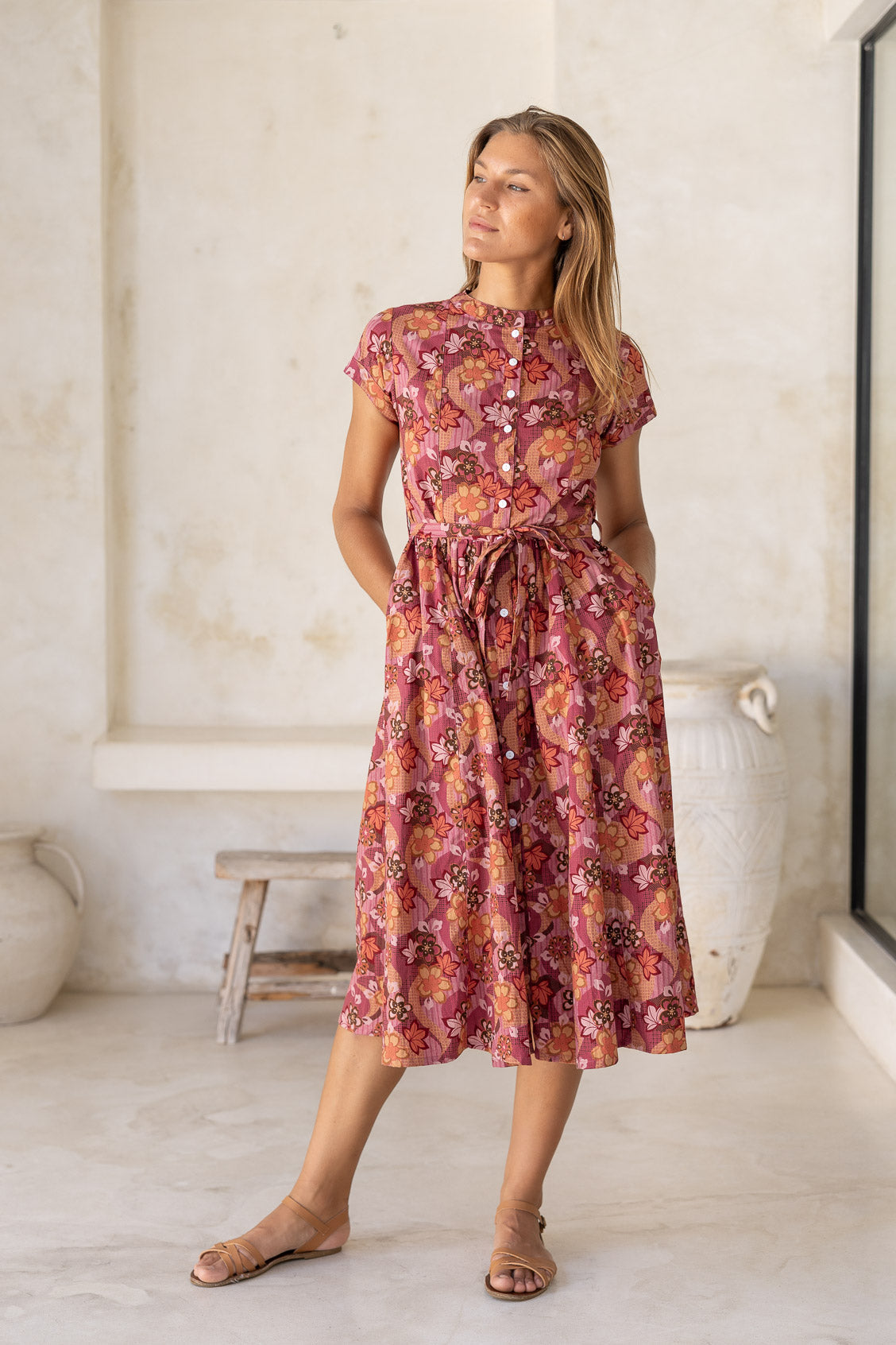 Woman standing with hands in pocket wears 100% cotton warm toned japanese style floral print dress with button through front and high round neckline.