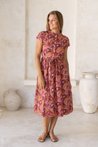Woman poses in retro style cotton button through dress with short sleeves, high round neckline and a fabric waist tie in a warm toned floral print.