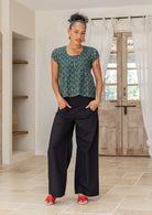 Woman wearing a green based floral print, 100% cotton top paired with a plain black pants standing in a minimalistic room.