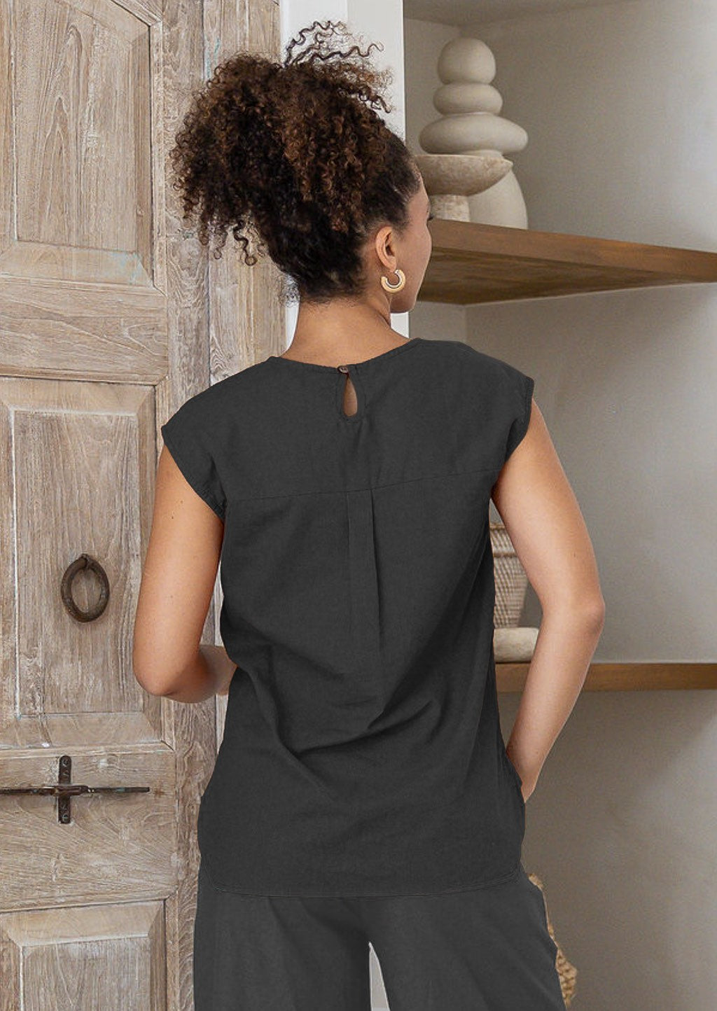 Woman wearing a black sleeveless cotton top with a keyhole and button clasp at the nape of the neck and a central box pleat 