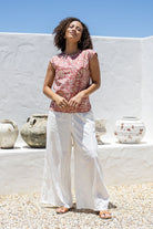 Woman wearing a warm pink based floral cotton top and white pants standing in front of a white wall