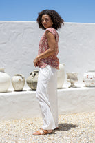 Woman in a warm pink based floral sleeveless cotton top and white pants standing in front of a white wall