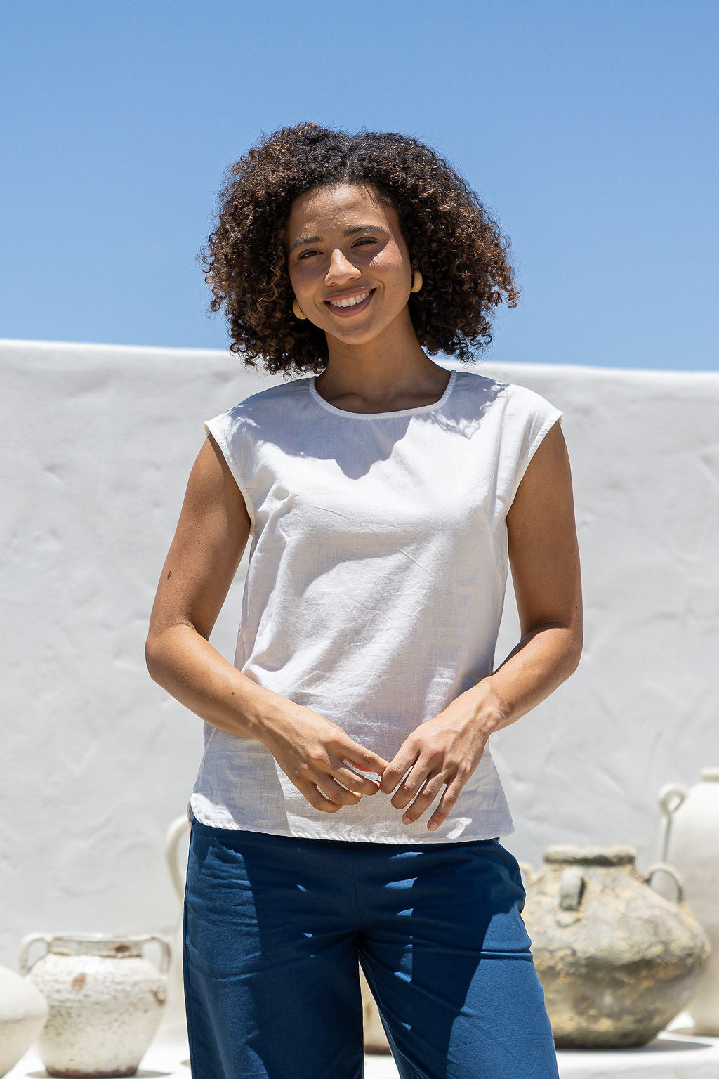 Woman wearing a white sleeveless cotton top and blue pants standing outdoors with a clear blue sky.