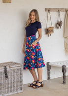 Woman standing sideways wearing a basic top and royal blue based floral cotton skirt in a home setting