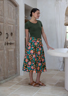 Woman standing sideways wearing a basic top and olive green based floral cotton skirt in a home setting