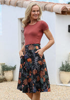 Woman wearing a basic rust top and black floral a-line cotton skirt with side zipper and both hands in pocket.