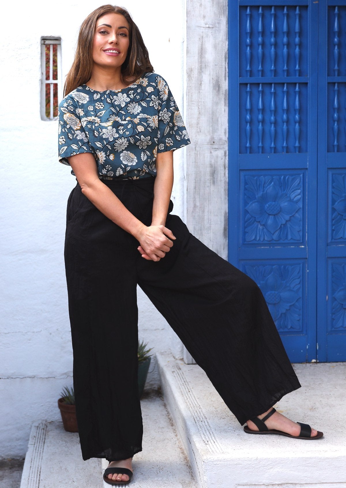 Woman poses on steps in black wide leg cotton pants paired with a blue floral top