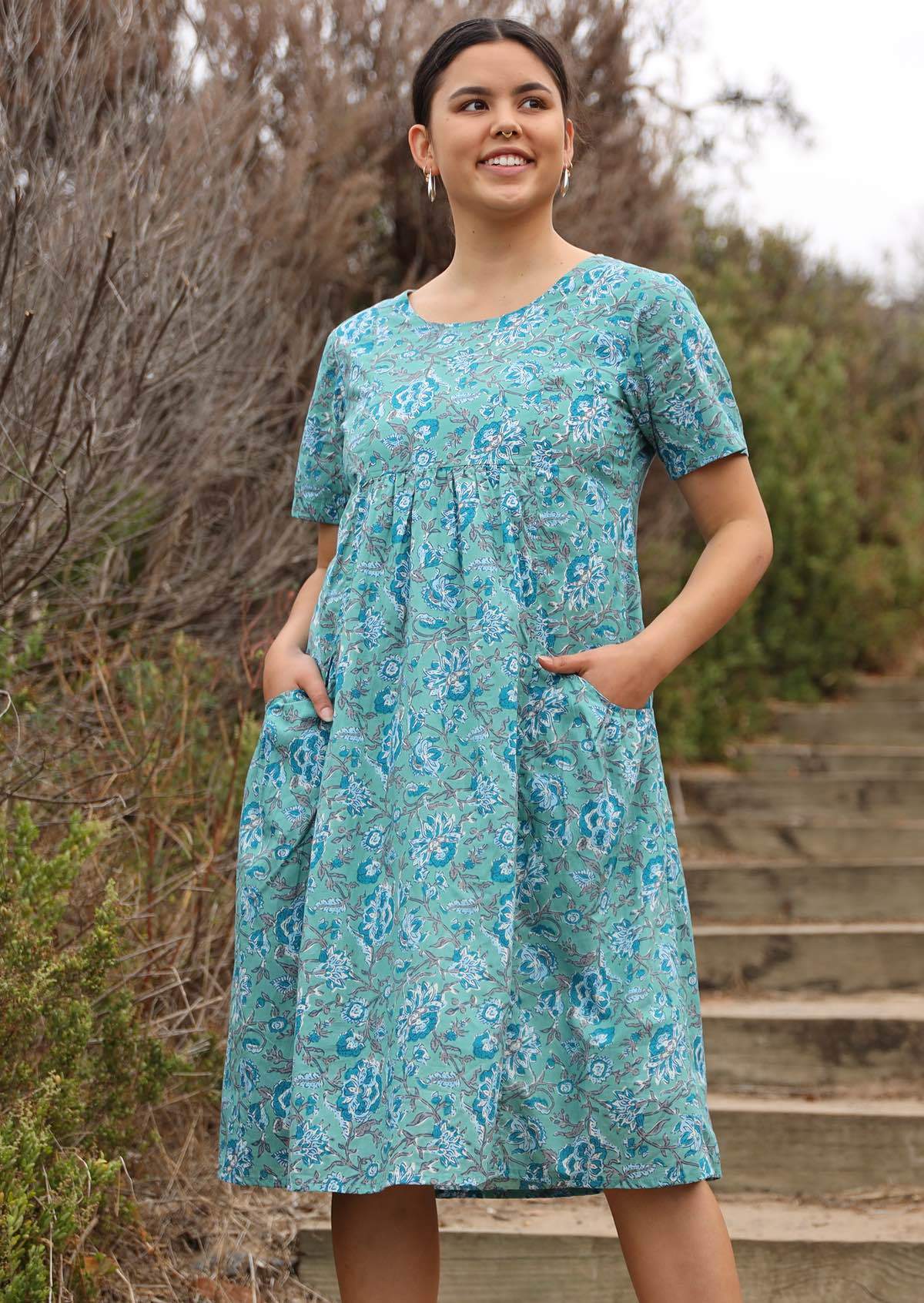 Model wears floral printed cotton dress in aqua shade with short sleeves and hands in pocket.