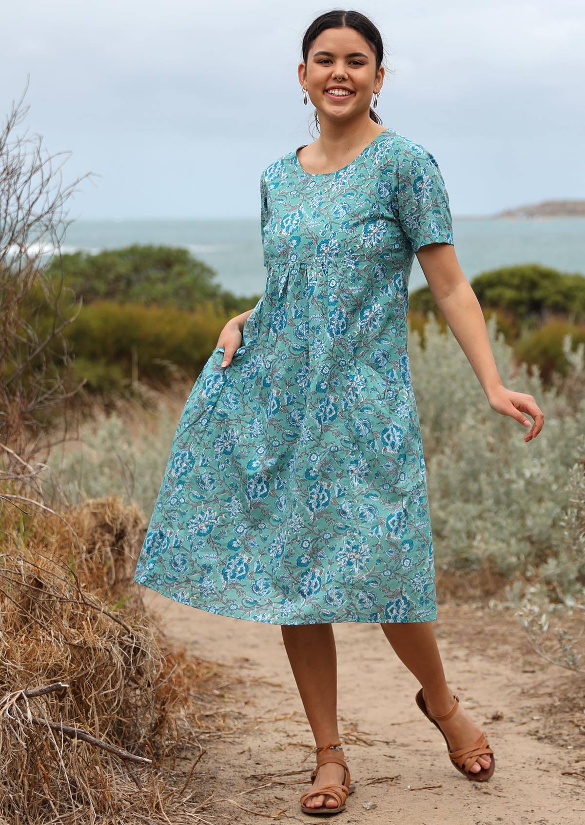 Model posing in knee length, 100% cotton dress in Indian floral print on aqua blue base.