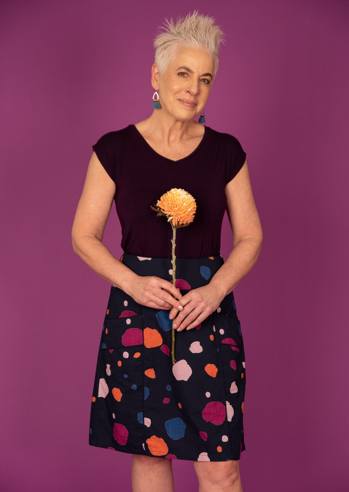 Woman holds flower in front of blue based knee length skirt with blue, orange, plum and pale pink splotch print, paired with a dark purple top