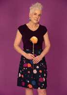 Woman holds flower in front of blue based knee length skirt with blue, orange, plum and pale pink splotch print, paired with a dark purple top