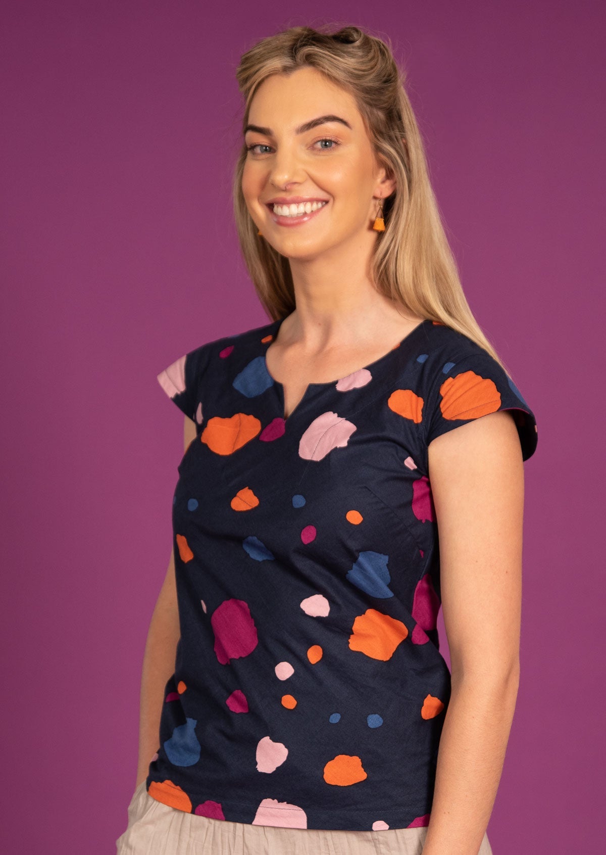 Woman wears a round neck key hole top with orange, blue, pink and plum spotty print an a navy blue base
