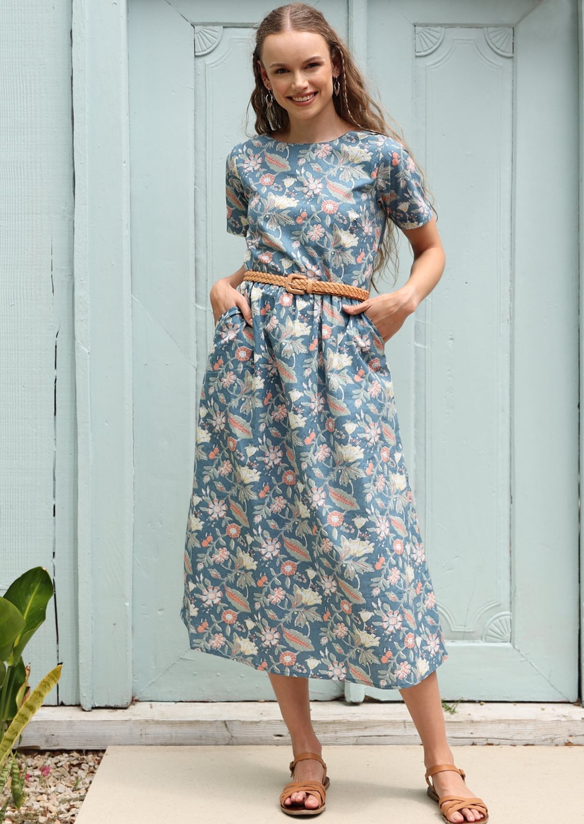 Woman poses in cotton midi length dress in a floral print with a mid blue base, paired with a plaited belt, with her hands in pockets