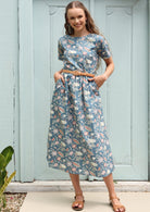 Woman poses in cotton midi length dress in a floral print with a mid blue base, paired with a plaited belt, with her hands in pockets