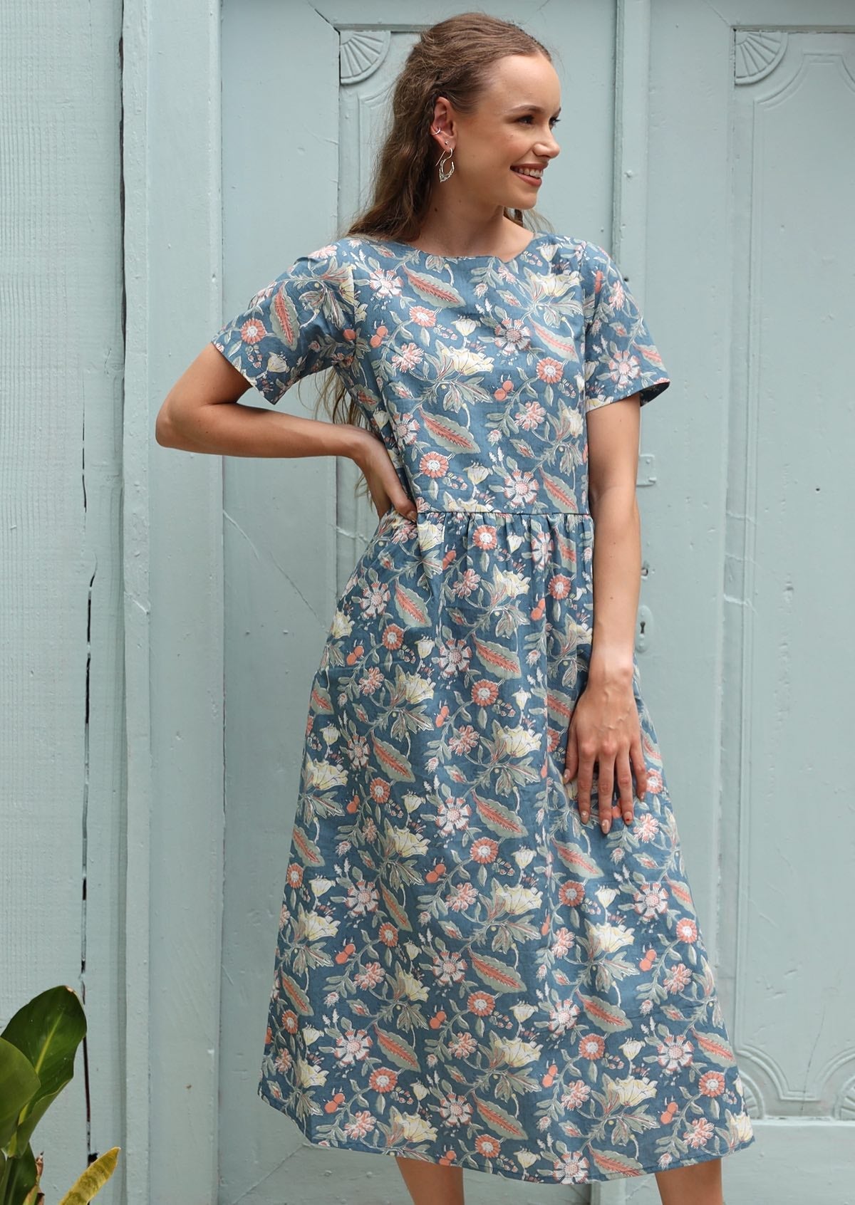 Woman wears midi length cotton dress with short sleeves, in coral, sage and lemon floral print on a mid blue base