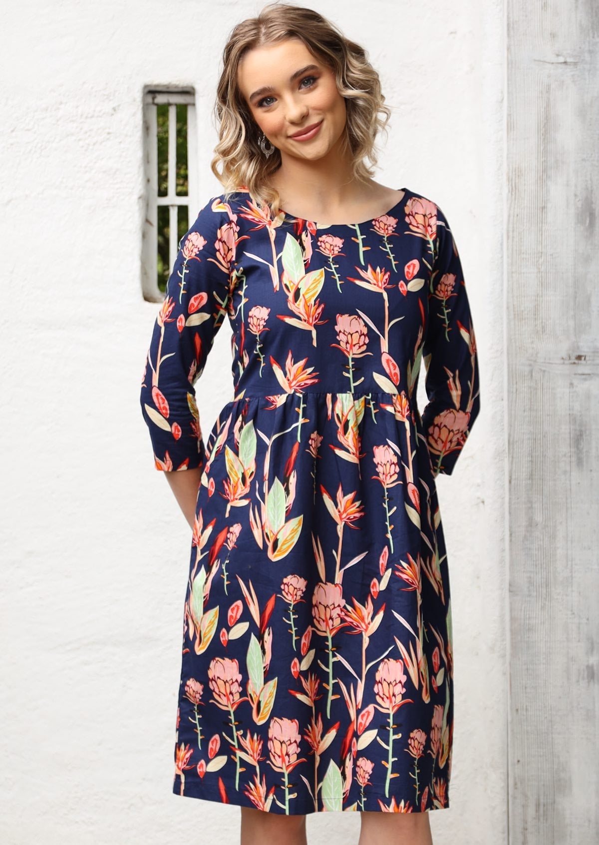 Woman wears wide round neck, 3/4 sleeve, knee length dress with pink and green toned native botanic print