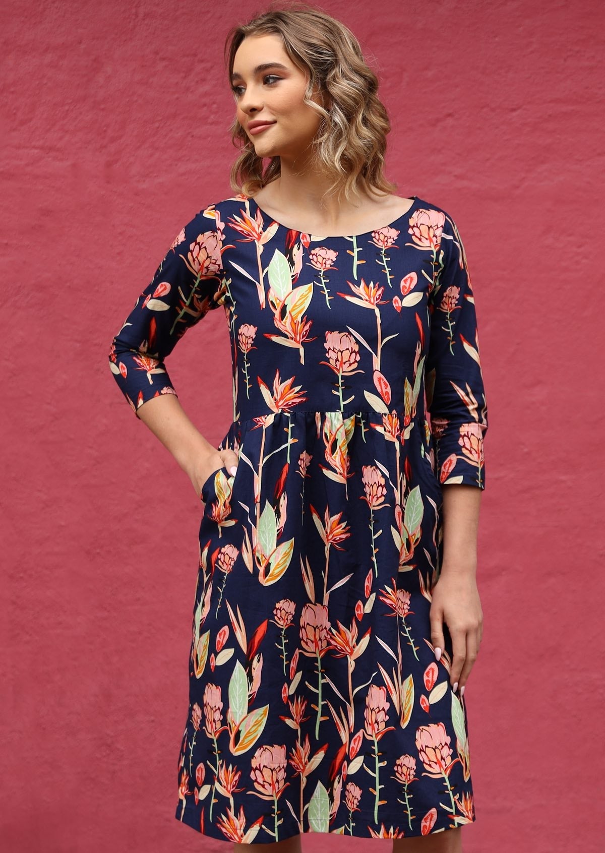 Woman wears knee length dress with 3/4 sleeves and pockets in navy based pink floral print.